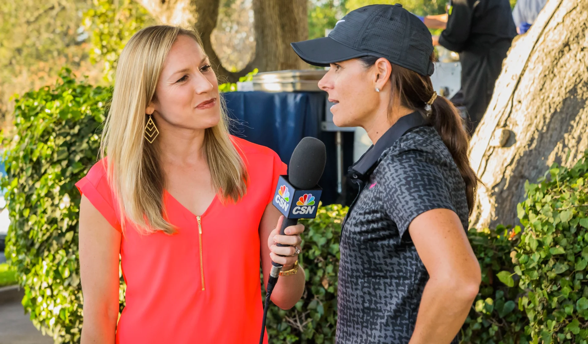 A woman in a coral blouse interviews another woman in a cap outdoors.