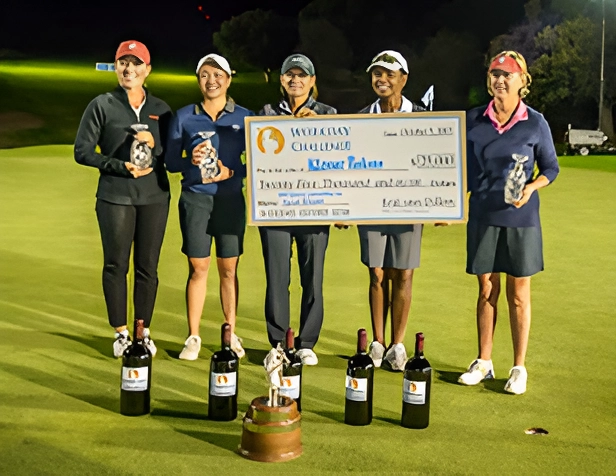Five women holding trophies and a large cheque on a grassy field at night.