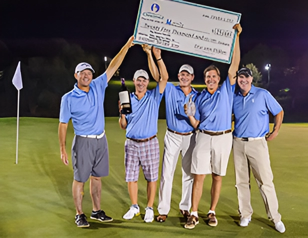 Five men in blue shirts holding a giant check on a golf course.